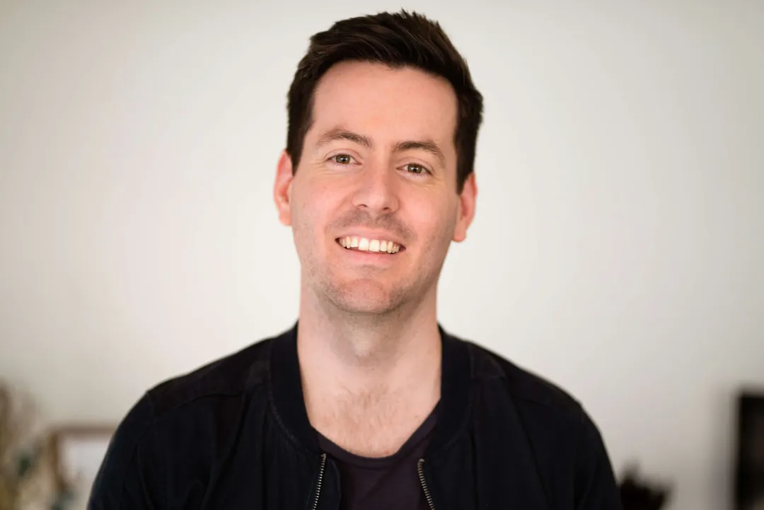 A portrait of a smiling man in a dark jacket against a plain wall