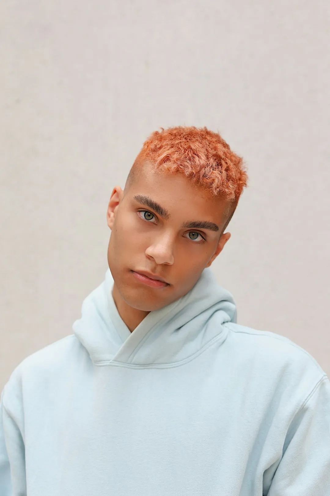 A young man with orange hair and a light blue hoodie in front of a plain light wall